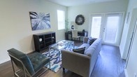 This single-floor space offers a thoughtfully designed living area that balances comfort and functionality. The living room is the centerpiece of the home, featuring rich hardwood flooring that runs throughout and large windows dressed with white blinds that invite generous amounts of natural light, creating a warm and welcoming ambiance. The space is styled with a neutral-toned sofa complemented by patterned accent pillows, while a bold blue and white geometric area rug anchors the seating area and adds a vibrant pop of color. A sleek glass-top coffee table with a chrome frame serves as a contemporary focal point, paired with a tufted green leather accent chair for added visual interest. A dark wood open-shelf bookcase lines one wall, providing both storage and display space, while a decorative starburst wall clock adds an artistic touch. Tucked neatly near the rear, a compact desk and chair set creates a functional workspace bathed in light from the adjacent door, which opens to offer easy access to the exterior. A curated wall art piece depicting a canopy of trees adds a nature-inspired element, reinforcing the airy and relaxed character of the overall space.