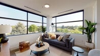 Located at 309 E 15th St in Vancouver, Washington, this contemporary single-floor residence showcases a thoughtfully designed open layout with clean lines and high-end finishes throughout. The living area is defined by expansive floor-to-ceiling windows on two walls, flooding the space with natural light and offering panoramic treetop views of the surrounding neighborhood. The modern kitchen is a standout feature, equipped with stainless steel appliances, white cabinetry, a large island with a matte black faucet, and generous countertops, all anchored by large-format polished tile flooring and a wide window overlooking the greenery beyond. A sleek hallway with polished concrete floors connects the main living areas to the private quarters. The bedroom is comfortably sized, with a large black-framed window that continues the theme of bringing the outdoors in. Two well-appointed bathrooms feature floating vanities, backlit mirrors, warm stone-toned tile flooring, and clean white walls. The overall ambiance of the residence is modern, bright, and airy, combining minimalist architectural design with warm, inviting interiors for an elevated living experience.