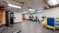 Located at 111 W Lamme St in Bozeman, Montana, this fitness center provides a comprehensive and well-organized workout facility within a compact, efficiently designed single-floor layout. The space features durable speckled rubber flooring throughout, ideal for high-traffic exercise use, and is illuminated by recessed panel lighting that ensures a bright, consistent environment.

The main workout area is divided into distinct functional zones. A cardio section lines one wall with multiple treadmills, an elliptical trainer, and a stationary cycle, offering a solid range of aerobic equipment. Adjacent to this, a strength training zone features a full dumbbell rack with a wide range of weights, a flat workout bench, a cable resistance machine, and a large wall mirror that enhances the sense of space and supports proper form during exercise. Stability balls and other functional training accessories are also available.

Bold orange accent walls add a vibrant, energizing aesthetic to the space, contrasting with the neutral grey walls and exposed ceiling infrastructure that gives the facility an open, industrial character. A corridor leads to an ancillary room with access to additional amenities, including what appears to be restroom facilities. A window along one wall allows natural light to filter in, complementing the overhead lighting. Overall, the fitness center is a practical, well-appointed space suited for a variety of workout routines.