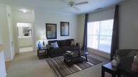This single-floor residence offers a welcoming and functional living environment designed for everyday comfort. The living room serves as the heart of the space, featuring plush carpeted flooring, neutral wall tones, and a ceiling fan that contributes to a relaxed, airy atmosphere. A large window dressed with elegant drape panels floods the room with generous natural light, creating a bright and inviting ambiance. The living area is thoughtfully arranged with ample room for seating and a central coffee table, making it ideal for both relaxing and entertaining. Decorative framed artwork adorns the walls, adding a touch of personality and warmth to the space. A side table with a stylish accent lamp provides soft supplemental lighting, enhancing the cozy feel of the room. The open sightlines from the living area extend toward adjacent rooms, suggesting a well-connected and easy-to-navigate floor plan. Overall, this single-story residence balances practicality with comfort, offering a clean, move-in-ready interior with tasteful finishing touches throughout.