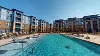 Located at 6026 Station Crossing Ave in Charlotte, NC, this outdoor amenity space offers a resort-style experience designed for relaxation and recreation. The centerpiece is a generously sized swimming pool with crystal-clear turquoise water, complemented by built-in aquatic lounge chairs and decorative water jet features that create a lively, refreshing atmosphere. The pool area is framed by a spacious concrete and paver deck, lined with rows of chaise lounge chairs ideal for sunbathing under the Carolina sky. A shaded pergola structure anchors a secondary seating and social area, furnished with tables and chairs for outdoor dining or gathering. Umbrella-shaded loungers provide additional comfort throughout the deck. The entire pool area is enclosed by a black iron security fence with a clearly posted pool rules sign at the entrance, ensuring a safe and well-managed environment. The surrounding multi-story residential buildings, clad in a combination of brick and modern panel siding with private balconies, create a cohesive courtyard setting that enhances the sense of community. A dedicated clubhouse structure with large windows and a covered porch is visible poolside, offering an indoor-outdoor connection. Manicured landscaping with young trees adds a finishing touch to this well-appointed outdoor retreat.