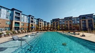 Located at 6026 Station Crossing Ave in Charlotte, NC, this outdoor amenity space offers a resort-style experience designed for relaxation and recreation. The centerpiece is a generously sized swimming pool with crystal-clear turquoise water, complemented by built-in aquatic lounge chairs and decorative water jet features that create a lively, refreshing atmosphere. The pool area is framed by a spacious concrete and paver deck, lined with rows of chaise lounge chairs ideal for sunbathing under the Carolina sky. A shaded pergola structure anchors a secondary seating and social area, furnished with tables and chairs for outdoor dining or gathering. Umbrella-shaded loungers provide additional comfort throughout the deck. The entire pool area is enclosed by a black iron security fence with a clearly posted pool rules sign at the entrance, ensuring a safe and well-managed environment. The surrounding multi-story residential buildings, clad in a combination of brick and modern panel siding with private balconies, create a cohesive courtyard setting that enhances the sense of community. A dedicated clubhouse structure with large windows and a covered porch is visible poolside, offering an indoor-outdoor connection. Manicured landscaping with young trees adds a finishing touch to this well-appointed outdoor retreat.