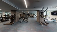This fitness center is a thoughtfully designed, single-floor facility offering a full suite of exercise equipment within a spacious and open layout. The space is anchored by patterned, multi-toned carpeted flooring that runs throughout, complemented by clean white and warm taupe walls accented by structural columns clad in textured stone-like finishes.

Overhead, linear LED lighting runs the length of the ceiling, providing consistent, bright illumination across the entire floor. The cardio zone features a row of treadmills and additional cardio machines positioned in front of large, floor-to-ceiling windows that frame views of lush tropical landscaping and the surrounding outdoor environment, filling the area with natural light and an open, airy feel.

The strength training section is well-appointed with a variety of selectorized cable and weight machines, including functional trainers, bench press stations, and other resistance equipment — all from professional-grade brands. Wall-mounted flat-screen televisions are positioned throughout the space for entertainment during workouts, and full-length mirrors enhance the sense of depth and allow for form-checking.

Entry to the fitness center is through a set of glass double doors, creating a clean and professional transition. The overall ambiance is modern and functional, striking a balance between an inviting atmosphere and a serious training environment.