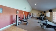 Located at 1651 S Dobson Rd in Mesa, Arizona, this single-floor fitness center is thoughtfully designed to support a well-rounded workout experience. The main exercise area is characterized by dramatic vaulted ceilings that rise to a striking peak, creating an open and airy atmosphere. Bold terracotta-red accent walls contrast with neutral tones, adding energy and visual interest to the space.

The strength training section is well-equipped with a variety of selectorized weight machines, including cable systems, leg press, and seated resistance equipment, all arranged along mirrored walls that enhance the sense of space and allow users to monitor their form. A full-length mirror spans one wall, amplifying the room's generous proportions.

The cardio zone is lined with large windows and glass-paneled doors that flood the area with natural light and offer views of the outdoor pool and surrounding landscape. Equipment includes treadmills, an elliptical trainer, and a stationary bike, all positioned to take advantage of the outdoor scenery. Wall-mounted televisions provide additional entertainment options.

A dedicated studio or stretching room features hardwood flooring, mirrored walls, natural light from large windows, and mounted screens, making it suitable for personal training, yoga, or guided workout sessions. Informational fitness posters are displayed throughout the facility, reinforcing a health-focused environment.