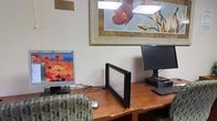This single-floor business center is thoughtfully designed to meet the practical needs of guests seeking a productive workspace. The room features multiple computer workstations arranged along a warm wood-toned desk surface, each equipped with monitors and keyboards to facilitate a range of tasks from browsing to document work. Comfortable upholstered seating with a classic patterned fabric is provided at the workstations, ensuring a relaxed experience during extended use. The space is finished with neutral wall tones that contribute to a calm, focused atmosphere. Decorative touches, including a framed floral artwork illuminated by a dedicated picture light above the desk area, add a welcoming warmth to what might otherwise feel like a purely utilitarian room. The overhead and accent lighting combine to keep the workspace well-lit and inviting. The layout is efficient, making good use of the available floor space while keeping the environment uncluttered. Overall, this business center offers a clean, organized, and comfortable setting well-suited for guests who need reliable access to computing resources during their visit.
