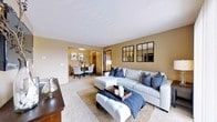 This single-floor residence offers a thoughtfully designed open-concept layout that seamlessly connects the living and dining areas, creating a cohesive and inviting atmosphere. The living room is a comfortable centerpiece, featuring plush wall-to-wall carpeting and warm, neutral-toned walls that lend a sense of warmth and sophistication. A generously sized sectional sofa in soft gray anchors the space, complemented by deep navy accent pillows and a tufted ottoman that doubles as a coffee table. Decorative grid-style mirrors adorn the walls, adding depth and visual interest to the room.

Natural light filters throughout, enhancing the open and airy feel of the space. A transition through a wide archway leads to the dining area, where a classic round table is surrounded by upholstered chairs beneath an elegant pendant light fixture, making it ideal for both casual meals and formal gatherings. Warm wood tones from the furniture and doorway accents provide a grounding contrast to the light carpet and neutral palette. The textured ceiling adds a subtle architectural detail that ties the rooms together. Overall, this residence combines functional design with tasteful styling to create a welcoming and livable environment.