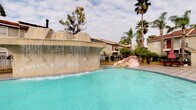 This community amenity space combines an inviting outdoor pool area with a well-appointed interior clubhouse. The centerpiece of the outdoor area is a generously sized swimming pool with crystal-clear turquoise water, highlighted by a striking stone waterfall feature that cascades dramatically into the pool. The surrounding landscape is enhanced by mature palm trees and lush greenery, creating a relaxed, resort-like atmosphere. A wrought iron perimeter fence and decorative boulders complete the outdoor setting.

The interior clubhouse features a grand, open lobby with soaring ceilings and a distinctive terrazzo floor showcasing an inlaid star pattern with contrasting dark tile borders. Rich wood paneling lines the walls, adding warmth and a classic character throughout. A traditional fireplace with an ornate wood mantle serves as a focal point in one area of the lobby. Natural light filters in through elegant French doors framed in dark wood molding, offering views of the outdoor courtyard. The space also accommodates a leasing or administrative area with workstations, as well as a fitness room visible through interior glass panels, making this clubhouse a fully functional and aesthetically refined community hub.