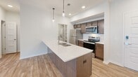This thoughtfully laid-out one-bedroom, one-bathroom residence offers a clean, modern aesthetic across its single floor. The open-concept kitchen and living area serves as the heart of the home, anchored by a large center island with a white quartz countertop, undermount sink, and stylish pendant lighting. Warm-toned cabinetry pairs with stainless steel appliances — including a refrigerator, electric range, and microwave — and a classic white subway tile backsplash adds a polished finishing touch. Light wood-look flooring flows throughout the main living area, enhancing the bright, airy feel created by recessed lighting and crisp white walls. The living room offers generous space and direct access to a private balcony through sliding glass doors, bringing in natural light and outdoor views. The bedroom is carpeted for comfort, featuring a window with a roller shade and a five-panel interior door leading to a dedicated closet. The bathroom is clean and functional, complete with a soaking tub and tiled shower surround, wood-look flooring, and a vanity with a light countertop. A separate laundry area houses a stacked washer and dryer unit, adding everyday convenience to this well-appointed residence.