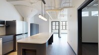 Located at 401 E State St in downtown Ithaca, NY, this 2-bedroom, 1.5-bathroom unit showcases a striking industrial-modern aesthetic throughout its well-appointed single floor. The open-concept living and kitchen area immediately impresses with soaring ceilings, exposed ductwork, and architectural curved column details that nod to the building's character. The kitchen is a standout, featuring a large center island with a fluted navy base and veined quartz countertop, sleek slate-gray cabinetry, stainless steel appliances including a French door refrigerator and dishwasher, a subway tile backsplash, and a globe pendant light cluster suspended overhead. Wide-plank hardwood flooring flows throughout the main living area, complementing the bright, airy atmosphere created by generous multi-pane, black-framed industrial windows that frame views of the surrounding Ithaca streetscape. The primary bedroom is a well-proportioned retreat bathed in natural light from a full bank of oversized windows. The bathrooms feature gray terrazzo-style tile flooring and clean, minimal finishes. Additional highlights include an in-unit washer/dryer, a dedicated closet, and a hallway that connects all spaces efficiently. The overall ambiance is contemporary and polished, blending loft-style industrial elements with modern residential comfort.