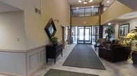 This lobby presents an impressive entryway characterized by dramatic double-height ceilings that lend a sense of grandeur to the space. The main entrance features a set of glass double doors framed in dark wood, allowing abundant natural light to flood the interior and offering views to the outdoors. Above the entrance, a clerestory window panel further enhances the brightness of the space.

The flooring consists of light-toned tile, complemented by a large, dark runner mat that extends from the entrance toward the interior, providing both practicality and visual definition. Wainscoting with panel detailing lines the lower portion of the walls, adding a refined, traditional character throughout.

A dark wood dresser with decorative hardware serves as a focal point along one wall, topped with a lush indoor plant and accented by a geometric diamond-shaped mirror above it. To the right, a seating area with upholstered chairs and a small side table creates a comfortable waiting or reception area. Recessed lighting and a chandelier overhead provide warm, even illumination, contributing to the lobby's polished and inviting ambiance.