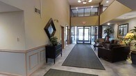 This lobby presents an impressive entryway characterized by dramatic double-height ceilings that lend a sense of grandeur to the space. The main entrance features a set of glass double doors framed in dark wood, allowing abundant natural light to flood the interior and offering views to the outdoors. Above the entrance, a clerestory window panel further enhances the brightness of the space.

The flooring consists of light-toned tile, complemented by a large, dark runner mat that extends from the entrance toward the interior, providing both practicality and visual definition. Wainscoting with panel detailing lines the lower portion of the walls, adding a refined, traditional character throughout.

A dark wood dresser with decorative hardware serves as a focal point along one wall, topped with a lush indoor plant and accented by a geometric diamond-shaped mirror above it. To the right, a seating area with upholstered chairs and a small side table creates a comfortable waiting or reception area. Recessed lighting and a chandelier overhead provide warm, even illumination, contributing to the lobby's polished and inviting ambiance.