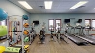 This fitness center offers a comprehensive workout environment designed to support a wide range of exercise routines. The space is laid out across a single floor and organized into distinct functional zones, allowing users to move seamlessly between cardio, strength, and functional training areas.

The cardio section features multiple treadmills, elliptical machines, and a stationary bike, each positioned near wall-mounted flat-screen televisions for entertainment during workouts. Natural light filters in through windows along one wall, complementing the bright, recessed panel lighting in the drop ceiling above.

A dedicated strength and functional training area houses a Matrix dual-cable functional trainer complete with a full attachment board, offering versatility for a wide variety of resistance exercises. Nearby, a rack of colorful medicine balls and stability balls provides options for dynamic and core-focused training, while kettlebells and exercise mats are available for floor work.

A large wall mirror spans one side of the space, enhancing the sense of openness and allowing users to monitor their form. Motivational phrases such as "No Pain No Gain" and "Eat. Sleep. Run." are displayed throughout, contributing to an energizing atmosphere. Patterned carpet flooring and light blue walls give the center a clean, welcoming aesthetic.