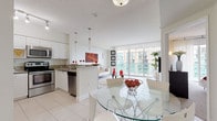Suite 2307 is a well-appointed high-rise residence offering a bright, open-concept floor plan finished with light tile flooring and a clean, contemporary aesthetic throughout. The kitchen is fully equipped with stainless steel appliances — including a range, microwave, dishwasher, and French door refrigerator — complemented by granite countertops, white cabinetry, and a peninsula that opens seamlessly to the dining area. A round glass dining table and modern seating anchor this space, creating a natural flow into the living room. The living area is generously proportioned, featuring large floor-to-ceiling sliding glass doors that flood the space with natural light and offer expansive views of the surrounding cityscape and neighboring buildings. The bedroom is a comfortable retreat, carpeted for warmth and outfitted with ample dresser storage, and also benefits from direct access to natural light via sliding glass doors. The bathroom is cleanly finished with tile flooring, a tub-shower combination, and granite-topped vanity. An in-unit washer and dryer provide added convenience. Throughout the suite, a cohesive palette of whites and neutrals creates a bright, airy ambiance well-suited to modern living.