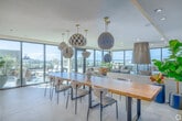 Located on Sixth Avenue in San Diego's vibrant Hillcrest neighborhood, this elevated amenity space offers a seamlessly integrated indoor-outdoor experience designed for both relaxation and entertaining. The interior opens with a sun-drenched lounge area dominated by floor-to-ceiling black-framed windows that frame sweeping panoramic views of the San Diego skyline and surrounding cityscape. A dramatic centerpiece freestanding brass fireplace with a cylindrical glass surround anchors the lounge, complemented by plush emerald velvet built-in seating and curated accent furnishings. Adjacent to the lounge, a generous dining area features a solid wood communal table beneath an artful cluster of woven pendant lights, creating an inviting atmosphere for gatherings. The stylish wet bar is finished with distinctive blue-green vertical tile, a curved quartz countertop, floating shelves, warm wood cabinetry, and bar seating — lending a polished, resort-like quality. A bright transitional sitting area with floor-to-ceiling glass connects seamlessly to the expansive outdoor terrace. Outside, the rooftop deck offers ample lounge seating around a fire pit table, a built-in stainless steel BBQ station surrounded by lush tropical plantings, and unobstructed open-sky views — all beneath the abundant San Diego sunshine.