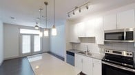 This single-floor space showcases a thoughtfully designed open-concept layout that seamlessly connects the kitchen and living areas. The kitchen is outfitted with clean white cabinetry extending to the ceiling, providing generous storage throughout. A white subway tile backsplash adds a classic yet contemporary touch, complementing the sleek countertops. Stainless steel appliances, including a built-in microwave and dishwasher, lend a modern, cohesive aesthetic to the space.

Overhead, a combination of track lighting and stylish pendant lights illuminates the kitchen and adjacent dining area, creating a warm, layered lighting scheme. The pendant fixtures, featuring brushed metal and glass designs, serve as subtle decorative focal points. A ceiling fan is visible in the adjoining living space, enhancing comfort and airflow.

The open layout allows natural light to filter through large windows at the far end of the space, brightening the interior and creating an airy atmosphere. The light-toned floors and neutral white walls further enhance the sense of openness. Overall, the space offers a clean, modern ambiance well-suited for everyday living.