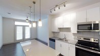 This single-floor space showcases a thoughtfully designed open-concept layout that seamlessly connects the kitchen and living areas. The kitchen is outfitted with clean white cabinetry extending to the ceiling, providing generous storage throughout. A white subway tile backsplash adds a classic yet contemporary touch, complementing the sleek countertops. Stainless steel appliances, including a built-in microwave and dishwasher, lend a modern, cohesive aesthetic to the space.

Overhead, a combination of track lighting and stylish pendant lights illuminates the kitchen and adjacent dining area, creating a warm, layered lighting scheme. The pendant fixtures, featuring brushed metal and glass designs, serve as subtle decorative focal points. A ceiling fan is visible in the adjoining living space, enhancing comfort and airflow.

The open layout allows natural light to filter through large windows at the far end of the space, brightening the interior and creating an airy atmosphere. The light-toned floors and neutral white walls further enhance the sense of openness. Overall, the space offers a clean, modern ambiance well-suited for everyday living.