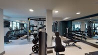 This modern fitness center is designed to support a comprehensive workout experience within a single, thoughtfully organized floor. Entry is made through double glass doors etched with a decorative leaf motif and "Fitness" branding, flanked by deep teal accent walls that set a contemporary tone throughout the space. The interior is divided into distinct functional zones, each tailored to a specific type of training. The cardio area lines the perimeter walls near expansive floor-to-ceiling windows, which flood the space with natural light and provide views of an outdoor amenity courtyard. A row of True-brand stationary bikes, ellipticals, and treadmills faces the windows, with wall-mounted televisions positioned for convenient viewing. The strength training area occupies the central and rear portions of the space, outfitted with a cable machine, free weight benches, adjustable racks, kettlebells, and a full dumbbell rack. Large wall mirrors extend the visual depth of the room and support proper form during exercise. A separate studio room, marked with its own branded glass doors and a patterned accent wall, provides additional space for group or individual training. Recessed lighting throughout ensures consistent illumination across all zones. The flooring transitions between durable light-toned rubber and carpet to delineate each area clearly.