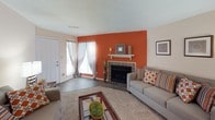 This single-floor residence offers a well-organized layout designed for comfortable everyday living. The living room serves as the centerpiece of the home, anchored by a tile-surround fireplace set against a bold orange accent wall, creating a warm and inviting focal point. Hardwood-style flooring runs through the main living area, complemented by natural light filtering through sheer curtains near the front entry.

Adjacent to the living room, the dining area features a ceiling fan with light fixture and connects seamlessly to the kitchen. The kitchen is outfitted with warm oak cabinetry, light countertops, a double-basin sink, and a full suite of appliances including an electric range with overhead ventilation.

The space includes two bedrooms, each finished with plush carpeting and textured ceilings. The primary bedroom features multiple windows that invite natural light, while the secondary bedroom includes a mirrored closet with vertical blinds. Both bedrooms offer generous space for furnishings.

Two full bathrooms serve the residence, each featuring a vanity with storage cabinetry, a large mirror, and a shower-tub combination. Built-in shelving in one bathroom adds additional storage utility. A dedicated laundry area with overhead shelving rounds out the functional amenities of this thoughtfully designed home.