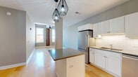 Located in the heart of downtown Buffalo on Main Street, this one-bedroom, one-bathroom unit offers a thoughtfully designed open-concept layout that seamlessly blends modern updates with industrial character. Light hardwood flooring runs throughout, complementing the soft gray walls and crisp white trim found across all rooms.

The combined kitchen and living area serves as the centerpiece of the unit. The kitchen is well-appointed with white shaker cabinetry, a herringbone-patterned white tile backsplash, dark countertops, and a full suite of stainless steel appliances including a refrigerator, range, microwave, and dishwasher. A central island with a dark countertop provides additional workspace and casual seating. Striking geometric pendant lights hang above, adding a contemporary industrial flair. The living area benefits from natural light through large windows framing a standout exposed brick column — a nod to the building's historic character — alongside sweeping views of the Buffalo skyline.

The bedroom is a comfortable, well-proportioned retreat with an arched window overlooking the city, hardwood floors, and access to a walk-in closet area. The bathroom features tile flooring, a soaking tub, and a vanity with white cabinetry and a dark countertop, maintaining the consistent modern aesthetic found throughout the unit.