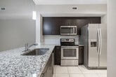 This 950-square-foot, single-story unit at 8881 SW 172nd Ave in Miami offers a practical and well-appointed layout across two bedrooms and two bathrooms. The open living area features light beige tile flooring and vaulted ceilings that lend a sense of spaciousness, with a sliding glass door opening onto a private balcony framed by lush tropical palm views. The kitchen is equipped with dark espresso cabinetry, granite countertops, a subway tile backsplash, and stainless steel appliances including a range, over-the-range microwave, and French door refrigerator. The primary bedroom measures 180 square feet and includes plush carpeting, a walk-in closet with wire shelving, and a dedicated en-suite bathroom featuring dark wood vanity cabinetry, granite countertops, and a glass-enclosed walk-in shower with tile surround. The second bedroom also features carpet flooring and a closet, with a window providing natural light. The second bathroom includes a tub/shower combination, tile surround, and matching dark vanity cabinetry. A dedicated in-unit laundry room with side-by-side washer and dryer completes the layout. Neutral warm-beige tones throughout create a cohesive, understated ambiance.