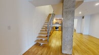Located at 1009 Rue De Bleury in the heart of Montréal, this two-level unit offers a well-designed layout spread across two floors. The main level features an open-concept living area and a fully equipped kitchen, where warm hardwood floors flow throughout. The kitchen is outfitted with wood cabinetry featuring frosted glass accents, granite countertops, a subway tile backsplash, and a full suite of stainless steel appliances including a dishwasher, range, and refrigerator. A striking open-tread staircase with a metal railing connects the two levels, framed by an exposed concrete structural column that lends an industrial character to the space. A powder room on the main level adds everyday convenience. The upper floor houses the spacious primary bedroom, which benefits from a window with city views and direct access to a full bathroom complete with a soaking tub, tiled surround, and a wood-finish vanity. Three closets — including a generous walk-in with wire shelving — provide ample storage throughout the unit. Clean white walls, consistent hardwood flooring across both levels, and the interplay of warm wood tones against raw concrete create a modern yet inviting urban aesthetic throughout this Montréal residence.