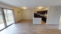 This single-floor unit at 140 Victor Dr in Hobart, Indiana offers a practical and well-organized layout. The open-concept living area is finished with light-toned hardwood-style flooring that extends throughout the space, creating a cohesive and clean aesthetic. A panel ceiling runs overhead, complemented by flush-mount lighting. The living area is equipped with a wall-mounted air conditioning unit and a baseboard heater, providing year-round climate control. A set of sliding glass doors opens to an outdoor patio area, allowing natural light to fill the room.

The kitchen is partially separated from the living space by a half-wall peninsula, providing both definition and an open feel. It features rich dark espresso cabinetry with ample storage, granite-style countertops, and black appliances including a coil-burner range with overhead ventilation hood, a refrigerator, and a sink with an exterior-facing window above it.

The bathroom is straightforward and functional, featuring a full tub-shower combination with a white surround, chrome fixtures, and a toilet. The overall layout of the unit is efficient and well-suited for comfortable single-floor living.