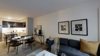 This single-floor space showcases a thoughtfully designed open-concept layout that seamlessly integrates the living, dining, and kitchen areas into one cohesive environment. The living area is anchored by a charcoal-toned sectional sofa accented with a draped throw blanket and flanked by a navy accent chair, creating a comfortable and stylish seating arrangement. Two framed abstract artworks mounted on the crisp white wall serve as a focal point, adding visual interest to the space.

The dining nook, positioned adjacent to the living area, features a round white table paired with sleek black chairs, making efficient use of the available space. A slim console table with metal legs bridges the two zones, styled with decorative objects that complement the overall aesthetic.

The kitchen, visible in the background, features clean-lined cabinetry in a light neutral tone with integrated appliances, maintaining the minimalist design language throughout. Light wood-toned flooring runs continuously across the space, enhancing the sense of openness and warmth. The smooth white ceiling and neutral walls contribute to a bright, airy ambiance, while carefully placed lighting adds depth and dimension to the overall atmosphere.