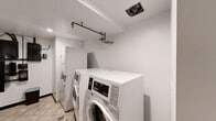 This single-room laundry facility is designed for practical, efficient use. The space features light-toned ceramic tile flooring throughout, providing a clean and durable surface suited to a high-use utility environment. The walls are finished in white, contributing to a bright and tidy atmosphere.

The room is equipped with multiple commercial-grade front-loading washing machines and stacked washer-dryer units, offering ample laundry capacity. The machines are digital and coin-operated, suggesting shared or communal use. Exposed overhead piping runs along the ceiling, serving the plumbing infrastructure of the units below, while a ceiling-mounted exhaust vent ensures proper ventilation within the space.

Along one wall, a bank of electrical panels, meters, and utility controls is mounted, indicating that the room also functions as a utility hub for the broader building. A white storage cabinet provides additional organizational space within the room.

The entry is accessed through a solid wood door with a brass knob. Recessed lighting fixtures provide consistent illumination throughout the room. The overall layout is straightforward and utilitarian, with every element oriented toward functionality. This laundry room serves as a well-equipped, centralized facility designed to meet the practical needs of its users efficiently.