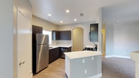This single-floor residence showcases a thoughtfully designed interior that blends modern aesthetics with functional living. The kitchen serves as the heart of the home, featuring rich dark cabinetry that contrasts beautifully against light countertops and neutral-toned walls. Stainless steel appliances, including a full-size refrigerator and a gas range, provide a contemporary and practical cooking environment. A central kitchen island offers additional prep space and adds to the open, airy feel of the layout. Recessed lighting throughout the kitchen ensures bright, even illumination, enhancing the clean lines of the space. Distinctive arched doorways add an elegant architectural detail, creating graceful transitions between rooms. Light hardwood-style flooring in the kitchen flows seamlessly toward carpeted living areas, adding warmth and texture underfoot. The open-concept design connects the kitchen to adjacent living spaces, promoting a sense of spaciousness and easy flow throughout the home. Natural light filters in through a window above the sink, contributing to the welcoming ambiance. The overall design is cohesive and modern, with neutral tones creating a versatile backdrop suited to a variety of interior styles.