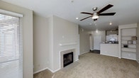 This single-story space in Addison offers an open, flowing layout that seamlessly connects the living area to the kitchen. The living room serves as the focal point of the home, anchored by a classic tile-surround fireplace with a white mantel, providing a warm and inviting atmosphere. Neutral-toned carpet covers the living area floor, contributing to the space's understated, comfortable aesthetic. A ceiling fan with dark wood-tone blades is centrally positioned overhead, adding both functionality and a subtle design element to the room.

The walls are finished in a soft, warm beige tone that complements the neutral palette throughout. Recessed lighting and pendant fixtures provide layered illumination across the open floor plan. The transition from the carpeted living area to darker flooring marks the entry into the kitchen zone, which features white cabinetry and a partial view of appliances. A door visible in the background suggests convenient access to additional areas of the home. Overall, the space presents a clean, move-in ready environment with a cohesive, neutral design scheme well-suited for a variety of furnishing styles.