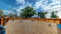 This outdoor dog park is a well-designed, fully enclosed recreational area built to provide dogs with a safe and stimulating environment. The ground is covered throughout with a generous layer of wood chip mulch, offering a comfortable and low-maintenance surface that is easy on paws and helps manage the overall upkeep of the space.

Enclosing the entire perimeter is a tall, sturdy cedar wood fence with a warm, natural finish, providing both security and a clean aesthetic. A gated entry point allows for controlled access, ensuring dogs remain safely within the space at all times.

The park is equipped with a variety of agility and enrichment features, including hoop jumps, a raised platform, and a balance bar, giving dogs opportunities for both physical activity and mental stimulation. Waste disposal stations are conveniently placed around the perimeter to support responsible pet ownership.

The open-air setting is bathed in natural light beneath a wide open sky, with mature trees visible beyond the fencing that provide partial shade and a pleasant natural backdrop. The surrounding urban context, including adjacent multi-story buildings, situates this park within a residential community setting.

Overall, this dog park combines practical amenities with a thoughtfully designed layout, making it an inviting and functional outdoor destination for pets and their owners.
