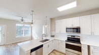 This single-floor residence showcases a clean, contemporary design with a cohesive neutral color palette and wood-look flooring that flows throughout the space. The kitchen is a standout feature, outfitted with crisp white shaker-style cabinetry extending to the ceiling for generous storage, quartz countertops, a classic white subway tile backsplash, and a full suite of stainless steel appliances including a range, built-in microwave, dishwasher, and refrigerator. A skylight overhead floods the kitchen with natural light, while stylish pendant lights hang above the peninsula, which includes an undermount sink. The open layout connects the kitchen seamlessly to the living area, which features a ceiling fan and natural light from a large window with blinds. The bedrooms are well-proportioned, each equipped with ceiling fans, wood-look flooring, and windows that invite natural light. The bathrooms are finished with white vanities, quartz countertops, large mirrors, and tile-surround showers. One bathroom includes a double-door closet for added storage convenience. Throughout the unit, the design emphasizes light, openness, and modern functionality.