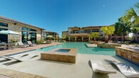 Located at Bella Terra Center Way in Richmond, Texas, this outdoor amenity space showcases a resort-inspired design centered around a generously sized swimming pool with crystal-clear water. The pool features a distinctive wading shelf with in-water sun loungers, allowing residents to relax directly in the shallow water. An adjacent raised spa with stone surround complements the main pool, offering a dedicated area for hydrotherapy and relaxation.

The surrounding pool deck is finished with decorative concrete and brick-inlaid pavers, providing ample space for lounge chairs and outdoor furniture. Shaded gathering areas with umbrella-covered tables offer comfortable spots for socializing or dining outdoors. A covered cabana pavilion with stone columns and a tile roof provides additional shade and houses outdoor grilling stations, ideal for entertaining.

The landscaping features a curated mix of palm trees, shade trees, and manicured greenery that enhances the resort atmosphere. A separate lawn area adjacent to the pool deck includes recreational options such as cornhole, creating a casual activity zone. The entire space is enclosed by a wrought iron fence with a secure gated entry. The architecture of the surrounding multi-story buildings features warm stucco and natural stone facades, tying the outdoor space cohesively into the broader Mediterranean-style community aesthetic.
