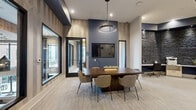 Located in Centennial, CO, the Sky Deck is a thoughtfully designed single-floor lounge space that blends modern aesthetics with warm, natural materials. The open living area is anchored by a striking dark tile fireplace wall, complete with a linear electric fireplace and a wall-mounted TV above a natural wood mantle. Flanking sconce lighting adds a warm, ambient glow to the space. A second mounted TV is positioned along a complementary wood-paneled accent wall, offering flexible viewing throughout the room. Seating is varied and well-appointed, featuring a rich cognac leather sectional sofa, sleek lounge chairs, and a dedicated dining area with leather chairs around a round table. A bar-height counter with dark wood stools and natural tree stump accents provides casual seating and workspace. The flooring throughout is a light-toned terrazzo-style tile, lending the space a clean, polished feel. Glass partition walls and frosted-glass doors define semi-private nook areas, allowing natural light to filter through while maintaining a sense of openness. Large French doors open to an outdoor deck, framing views of the surrounding Centennial skyline. The overall ambiance is contemporary yet inviting, blending urban sophistication with natural warmth.