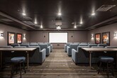 Located at 31611 Pete Von Reichbauer Way S in Federal Way, Washington, this private screening theater delivers an immersive cinematic experience in a thoughtfully designed single-level space. The main screening room is anchored by a large front projection screen paired with a ceiling-mounted projector, providing a true theater-quality viewing setup. Rows of generously sized, gray upholstered lounge chairs with built-in cup holders are arranged in a tiered layout along a central aisle, ensuring clear sightlines from every seat. The dark, richly toned ceiling and warm wall sconces create an intimate, dimly lit ambiance reminiscent of a classic cinema. Patterned carpet in earthy tones of brown, gray, and teal runs throughout, adding texture and visual interest underfoot. Classic framed movie posters — including iconic titles — adorn the walls, reinforcing the cinematic theme. A secondary area adjacent to the main room features a countertop workspace with cabinetry, bar-height seating, and additional movie memorabilia, ideal for pre-show gatherings or refreshments. Bold red double doors with porthole windows serve as the dramatic main entrance, completing the theatrical aesthetic. Recessed ceiling lighting and wall-mounted sconces provide layered illumination throughout the space.