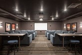 Located at 31611 Pete Von Reichbauer Way S in Federal Way, Washington, this private screening theater delivers an immersive cinematic experience in a thoughtfully designed single-level space. The main screening room is anchored by a large front projection screen paired with a ceiling-mounted projector, providing a true theater-quality viewing setup. Rows of generously sized, gray upholstered lounge chairs with built-in cup holders are arranged in a tiered layout along a central aisle, ensuring clear sightlines from every seat. The dark, richly toned ceiling and warm wall sconces create an intimate, dimly lit ambiance reminiscent of a classic cinema. Patterned carpet in earthy tones of brown, gray, and teal runs throughout, adding texture and visual interest underfoot. Classic framed movie posters — including iconic titles — adorn the walls, reinforcing the cinematic theme. A secondary area adjacent to the main room features a countertop workspace with cabinetry, bar-height seating, and additional movie memorabilia, ideal for pre-show gatherings or refreshments. Bold red double doors with porthole windows serve as the dramatic main entrance, completing the theatrical aesthetic. Recessed ceiling lighting and wall-mounted sconces provide layered illumination throughout the space.