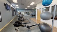 This fitness center is a spacious, single-floor facility designed to accommodate a wide range of workout activities. The layout is divided into distinct functional zones, providing dedicated areas for cardiovascular training, strength equipment, and open-floor exercise. The cardio section features a row of treadmills, stationary bikes, a rowing machine, and an elliptical trainer, all positioned along a wall lined with windows that allow natural light to fill the space and offer views of the surrounding outdoor environment. Wall-mounted televisions are strategically placed to keep users engaged during cardio sessions. The strength training area houses a multi-station cable machine along with additional weight machines and benches, supported by a large wall mirror that visually expands the space and aids in form monitoring. An open area with rubber flooring provides room for stretching, functional training, or use of the stability balls visible throughout the facility. The flooring transitions between textured rubber and hard-surface tile, delineating the workout zones clearly. The drop ceiling features recessed panel lighting that ensures consistent, even illumination across the entire floor. The walls are finished in a cool blue-gray tone, contributing to a focused and professional atmosphere. Restroom access and waste disposal stations are conveniently located within the facility.