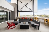 This contemporary amenity space seamlessly blends indoor comfort with open-air living across a single, generously sized floor. The architecture is defined by dramatic steel cross-bracing, exposed structural beams painted in deep navy, and soaring vaulted ceilings that amplify the sense of volume throughout. Large open-air sections dissolve the boundary between interior and exterior, flooding the space with natural light and offering sweeping panoramic views of the surrounding cityscape, palm trees, and distant hills.

The outdoor terrace area is anchored by a substantial L-shaped sectional sofa in charcoal grey, centered around a sleek rectangular fire pit table — an ideal setting for relaxed gatherings. Adjacent to the lounge, a dining zone features bold red chairs surrounding a white table, with a built-in counter and yellow bar stools lining the perimeter wall beneath the open sky. Lush snake plants in raised planters add greenery throughout.

The enclosed interior lounge offers a more intimate atmosphere, featuring a patterned geometric carpet, a minimalist daybed, vibrant yellow accent walls, wall-mounted planters, and open shelving. A split-system AC unit ensures year-round comfort. Together, the zones create a versatile, design-forward communal environment with a cohesive modern aesthetic.