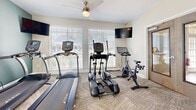This well-appointed fitness center at 1825 Grove Point Rd in Savannah, Georgia, offers a comprehensive workout environment across a single floor. The space is divided into distinct exercise zones, each designed to support a variety of fitness routines. The cardio area features treadmills, an elliptical trainer, and a stationary cycling bike, all positioned near large windows dressed with white blinds that invite ample natural light and outdoor views. Wall-mounted flat-screen televisions and ceiling fans enhance comfort during workouts. The strength and free weight zone is equipped with a multi-station cable machine, a full rack of dumbbells ranging in various sizes, and a weight bench, with large mirrors along the wall to aid in proper form. A dedicated functional training area stores stability balls in multiple sizes, resistance bands, and additional accessories on a vertical rack. The space is finished with neutral-toned patterned carpeting throughout, and the walls are painted in a calming combination of soft beige and sage green. A glass-panel door provides direct access to an outdoor area, adding convenience and ventilation. Towel dispensers, a waste receptacle, and posted fitness center guidelines reflect thoughtful amenities for users.