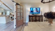 This single-floor amenity space is designed for entertainment and socializing, combining multiple functional zones within one cohesive, open layout. The interior is finished with warm hardwood-style flooring, crown molding, and neutral-toned walls accented by wainscoting, creating a polished yet welcoming atmosphere throughout.

The main lounge area serves as a home theater, anchored by a large projection screen and ceiling-mounted audio system. Comfortable seating, including a generous sofa and upholstered chairs, is arranged around a substantial wood-and-metal coffee table atop a chevron-patterned area rug. Decorative wall art and sculptural accents add visual interest.

Adjacent to the theater area, a billiards table occupies a generously sized open zone, complemented by additional seating arrangements with televisions mounted on the walls, allowing for multiple simultaneous activities.

A fully equipped kitchen features light wood cabinetry extending to the ceiling, marble-look countertops, a large island with bar seating, and a wall-mounted television. Geometric pendant lighting adds a contemporary touch overhead.

A quieter lounge nook near large windows offers natural light, leather seating, and a dining table with chairs, providing a more intimate setting within the broader space. Recessed lighting throughout ensures consistent illumination across all zones.