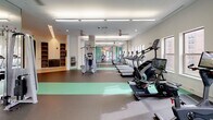 Located at 4700 Berwyn House Rd in College Park, Maryland, this single-floor fitness center offers a spacious, thoughtfully designed environment for a comprehensive workout experience. The facility is organized into distinct functional zones, seamlessly connected by an open, elongated floor plan with durable rubberized flooring in grey and green accents.

The cardio section features a long row of treadmills, ellipticals, stair climbers, and cycling machines — including Expresso interactive bikes — arranged to maximize space and airflow from ceiling fans overhead. Linear LED lighting runs throughout, complemented by recessed fixtures that keep the facility bright and energizing.

The strength training area is generously outfitted with a full dumbbell rack, multiple cable and resistance machines from brands such as Precor, along with benches and free weight stations. Floor-to-ceiling mirrors expand the sense of space and allow users to monitor their form. Large windows along the perimeter bring in natural light and provide views of the surrounding College Park neighborhood.

A separate spin or group exercise room is accessible through distinctive teal glass sliding doors, offering privacy and featuring globe pendant lighting for a distinct ambiance. Additional amenities include built-in shelving, wall-mounted TVs, and motivational fitness artwork displayed in the connecting corridors, creating a polished and functional athletic environment.