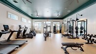 Located at 7100 Summerside Dr in Whitsett, NC, this fitness center offers a thoughtfully designed exercise space that caters to a wide range of workout needs. The room is defined by its striking teal-painted tray ceiling trimmed in deep green, lending a polished, upscale aesthetic that sets it apart from a typical gym environment. Multiple ceiling fans ensure comfortable air circulation throughout the space.

The flooring is a smooth, light-toned surface that keeps the area feeling open and clean. White beadboard wainscoting lines the lower walls, complementing the overall design and adding a classic touch. Natural light filters in through numerous windows positioned along multiple walls, creating a bright and welcoming atmosphere, while large glass French doors provide additional light and direct access to the outdoors.

The exercise room is comprehensively outfitted with cardio equipment including treadmills, elliptical machines, a stationary bike, and a rowing machine. The strength training area features a power rack with adjustable bench, a cable machine, a full dumbbell rack, and additional multi-function equipment. Wall-mounted mirrors with stylish gooseneck sconce lighting enhance the strength training zone. Two wall-mounted televisions provide entertainment, and built-in storage shelving adds functional convenience to this well-appointed fitness facility.