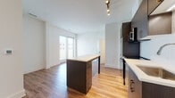 This contemporary single-floor residence showcases a thoughtfully designed open-concept layout that maximizes both space and functionality. The kitchen serves as the heart of the home, featuring rich dark-toned cabinetry paired with stainless steel appliances, including a refrigerator, microwave, and dishwasher. A classic white subway tile backsplash adds a clean, timeless touch, while a central island with a light quartz countertop provides ample workspace and casual dining potential. Track lighting overhead ensures the kitchen is well-illuminated at all times.

Warm wood-look flooring flows seamlessly throughout the unit, connecting each room with a cohesive and inviting aesthetic. The living area benefits from natural light streaming through sliding glass doors, creating a bright and airy atmosphere. The bedrooms are generously sized, each featuring large windows with blinds that allow for adjustable natural light and outdoor views.

The bathroom is finished with modern appointments, including a sleek dark vanity topped with a white quartz countertop, a backlit LED mirror, and a full tub-shower combination with clean white tile surround. Crisp white walls and light tile flooring complete the bathroom's clean, contemporary look. Overall, the space presents a polished, move-in ready environment with consistent modern finishes throughout.