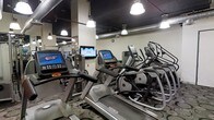 This fitness center offers a comprehensive workout environment designed to meet the needs of a wide range of users. The space is outfitted with a robust selection of modern cardio equipment, including Matrix-brand treadmills and elliptical trainers, each equipped with individual digital touchscreen displays for entertainment and workout tracking. Wall-mounted flat-screen televisions are positioned throughout the room, ensuring visibility from multiple workout stations. The ceiling features an exposed industrial aesthetic, with dark-painted ductwork and stylish pendant-style drop lights that provide ample illumination throughout the floor. The light-colored walls contrast with the darker ceiling, contributing to an open and energetic atmosphere. Large mirrors are mounted along the walls, enhancing the sense of space and allowing users to monitor their form during exercise. The patterned flooring adds a finished, professional look to the environment. The layout is thoughtfully organized to maximize the number of machines while maintaining comfortable spacing between equipment, allowing for ease of movement throughout the space. Overall, the fitness center presents a clean, well-maintained, and fully functional facility suitable for cardiovascular training.