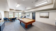This community game room is a thoughtfully designed recreational space offering a comfortable and inviting environment for social gatherings and leisure activities. The centerpiece of the room is a full-size pool table with a warm walnut finish and cream felt surface, complemented by a dedicated cue rack holding a full set of billiard cues. Recessed tray ceilings with flush-mounted rectangular light fixtures provide bright, even illumination throughout the space, while a clean white and neutral gray color palette creates a modern, polished atmosphere. Patterned gray carpet covers the floor, adding texture and a sense of warmth underfoot. Along one wall, a wall-mounted flat-screen television is paired with a bold navy-blue media console, creating a media viewing zone flanked by comfortable upholstered lounge chairs. A built-in wet bar with navy cabinetry and ample counter space offers a convenient refreshment station. On the opposite side of the room, a dining or game table with upholstered chairs provides additional seating. Windows along one wall allow natural light to filter in, offering views to the exterior. Framed landscape artwork adorns the walls, contributing to the room's refined yet relaxed ambiance. A dark-framed glass door provides access to an adjoining area or exterior corridor.