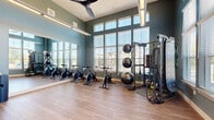 Located in Colorado Springs, CO, this fitness facility offers a thoughtfully designed exercise room that combines functionality with an inviting atmosphere. The space is characterized by soaring ceilings and an impressive bank of large, multi-pane windows that line the walls, flooding the room with natural light and providing open views of the surrounding neighborhood. Deep teal-green walls, white trim, and warm wood-look flooring create a modern yet energizing aesthetic throughout.

The exercise room is well-appointed with a row of indoor cycling bikes, a multi-station functional training rig, battle ropes, medicine ball storage, resistance bands, and a selection of free weights and accessories — catering to a wide range of workout styles. A large wall mirror expands the sense of space and allows for form checks during exercise. Ceiling fans and modern linear pendant lighting ensure comfort during intense workouts.

A dedicated open-floor area provides ample room for stretching, yoga, or bodyweight training, while a wall-mounted display screen enables guided workout sessions. The entrance features glass French doors and a stylish reception counter with marble-look surfaces and curated décor, setting a polished tone from the moment you arrive. This exercise room delivers a complete and comfortable fitness experience.
