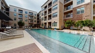 This expansive outdoor pool area is set within a beautifully designed courtyard, framed on multiple sides by a contemporary multi-story residential building featuring a blend of brick and stone facades with balconied units overlooking the space. The centerpiece is a generously sized swimming pool with crystal-clear blue water, complemented by built-in water features along one edge that add a tranquil, resort-like ambiance.

Surrounding the pool is a combination of warm-toned composite wood decking and smooth concrete patio surfaces, providing ample space for lounging and socializing. Comfortable chaise lounge chairs are thoughtfully arranged throughout the deck area, perfect for relaxing poolside.

A covered outdoor living area with a large shade canopy offers a sheltered retreat, furnished with additional seating, dining areas, and grilling stations — ideal for outdoor entertaining. Nearby, a more intimate seating nook features a long wooden bench, cylindrical accent stools, and lush manicured shrubbery that softens the hardscape with greenery.

Elegant arched architectural details, glass panel railings, decorative pendant lighting, and oversized statement planters add refined design touches throughout. The entire pool courtyard is enclosed within the building complex, creating a private and serene outdoor retreat.