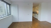 This single-floor space features a clean, minimalist design with warm hardwood-style flooring that runs throughout, creating a cohesive and inviting atmosphere. The living area is bright and open, with crisp white walls that enhance the sense of space and light. A sleek vertical panel radiator is mounted along one wall, serving as both a functional heating element and a subtle modern design feature.

The space flows naturally into the kitchen, which is defined by white cabinetry extending to the ceiling, providing ample storage. The kitchen is equipped with a white gas range, an under-cabinet range hood, and a dark countertop that offers a clean contrast against the white cabinetry. Recessed lighting in the kitchen ensures the workspace is well-lit. The layout allows for practical movement between the living and cooking areas, making it well-suited for everyday use.

Overall, the space presents a tidy, contemporary aesthetic with a neutral color palette. The combination of warm flooring tones and bright white finishes gives the unit a fresh, modern feel throughout.