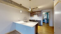 Located at 213 Belmont Ave E in Seattle's vibrant Capitol Hill neighborhood, this two-level loft-style unit blends industrial character with modern finishes throughout. The main floor opens with a dramatic living area featuring soaring ceilings, light wood flooring, and floor-to-ceiling glass entry doors that flood the space with natural light and offer views of the surrounding streetscape. Exposed concrete columns and ceiling beams add a striking industrial aesthetic that carries through the entire level.

The open-plan kitchen and living area anchor the main floor. The kitchen is outfitted with stainless steel appliances, including a range and refrigerator, dark wood cabinetry, white tile backsplash, and a generous white-countertop island with an undermount sink and pendant lighting overhead. Track lighting complements the space's warm, functional ambiance.

An open-tread wood staircase with a metal railing leads to the upper level, which houses the second living area, two bathrooms, and a spacious walk-in closet. Both bathrooms feature dark stone-finish countertops, modern vanities with ample storage, wood-look flooring, and clean white tile. The walk-in closet includes built-in wire shelving systems for organized storage. Throughout both floors, the consistent use of wood flooring, neutral tones, and industrial accents creates a cohesive and contemporary living environment.