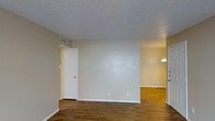 This single-floor space presents a clean, straightforward layout designed for functional living. The interior is finished with warm-toned hardwood floors that extend throughout the visible areas, providing a cohesive look across the open floor plan. Neutral beige walls create a blank canvas, making the space adaptable to a variety of décor styles.

The main living area flows openly into an adjacent room, visible in the background and illuminated by a wall-mounted light fixture, suggesting a dining or secondary living area. A white door on the left side of the main room leads to what appears to be a closet or utility space, adding practical storage to the layout.

The textured popcorn ceiling adds a classic architectural detail while housing a centrally placed HVAC vent, ensuring climate control throughout the space. White baseboards trim the perimeter of the rooms, offering a clean contrast against the hardwood floors and neutral walls.

Overall, the space maintains a simple, well-maintained aesthetic. Its open connectivity between rooms, consistent flooring, and neutral color palette make it a versatile environment suitable for a range of uses.